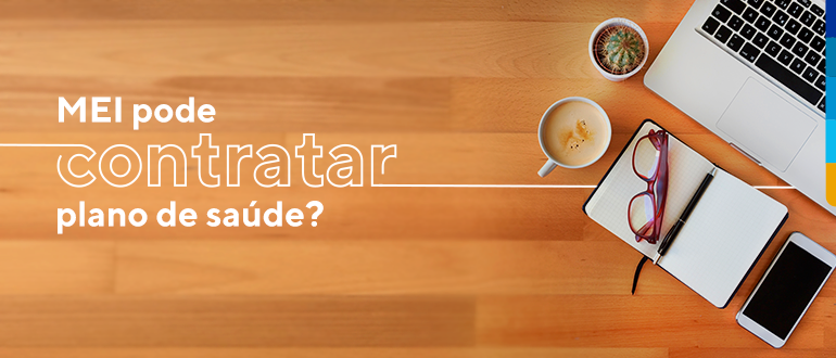 Texto: MEI pode contratar plano de saúde?, sobre fundo da madeira. Ao lado computador, celular, caderneta de anotação, xicara de café e um mini cactos sobre a mesa.