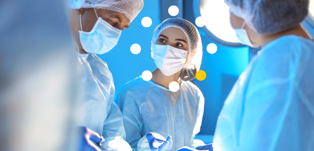 Imagem de 3 pessoas com máscaras, touca, luvas e avental de centro cirúrgico.