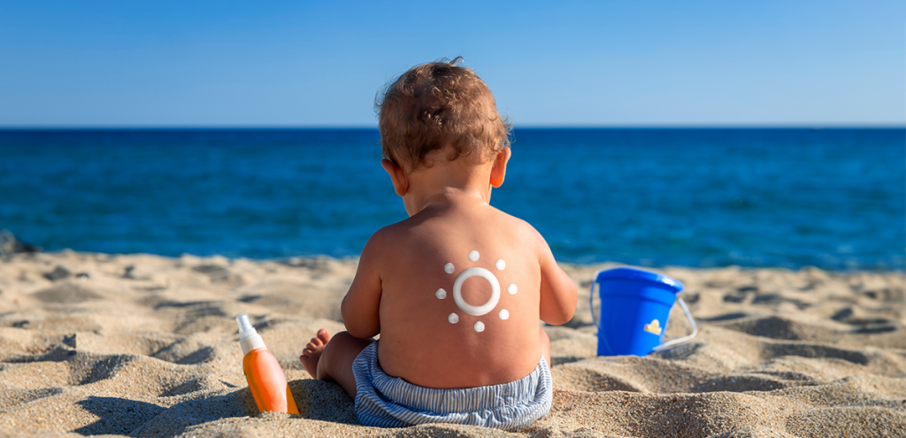 Criança sentada na areia da praia de frente ao mar, com um baldinho e o protetor solar ao lado. Simbolizando Dezembro Laranja, a prevenção contra o contra o Câncer de pele