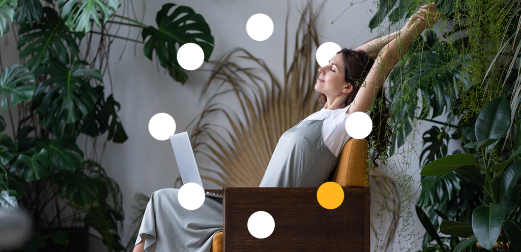 imagem de uma mulher sentada em uma poltrona com um notebook no colo, em um ambiente cheio de plantas, simbolizando equilíbrio entre vida pessoal e profissional.