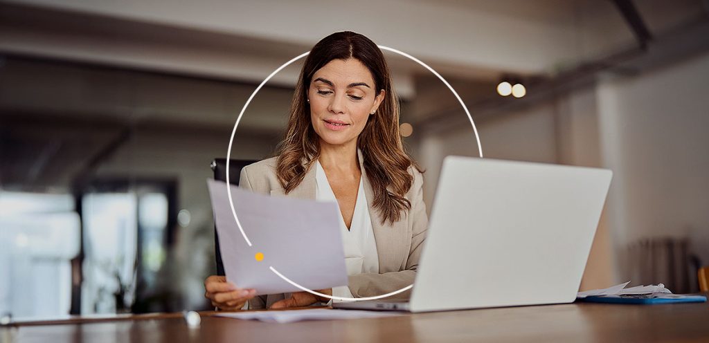 mulher sentada na frente de um notebook, segurando uma folha e uma das mãos, simbolizando IR 2024