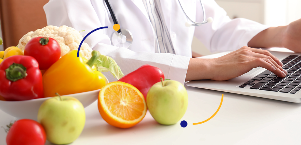 Imagem de duas mãos digitando no notebook com alimentos do lado, simbolizando uma nutricionista