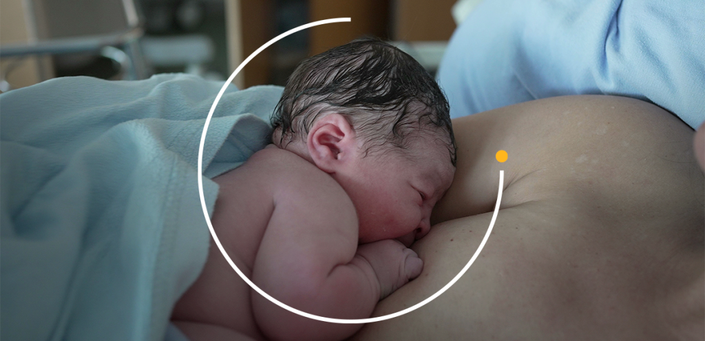 Foto em tom quente e iluminação natural de uma mãe em contato pele a pele com o recém-nascido no tórax logo após o parto, transmitindo acolhimento e proteção